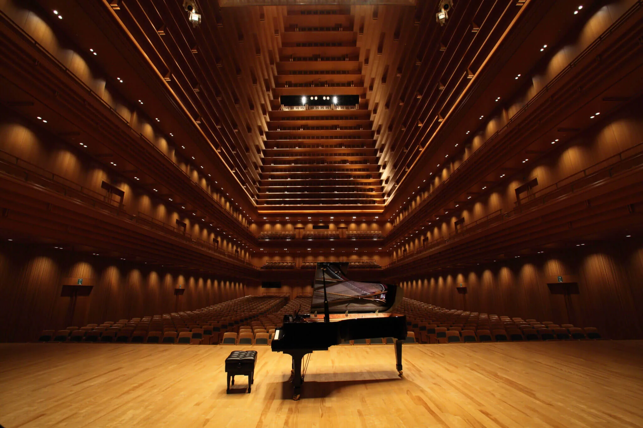 A black grand piano alone on the stage of a wood-panelled concert hall